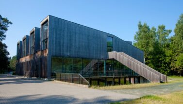 Modern building made primarily of wood and glass is in a green area, perhaps a forest. The weather is nice and sunny.