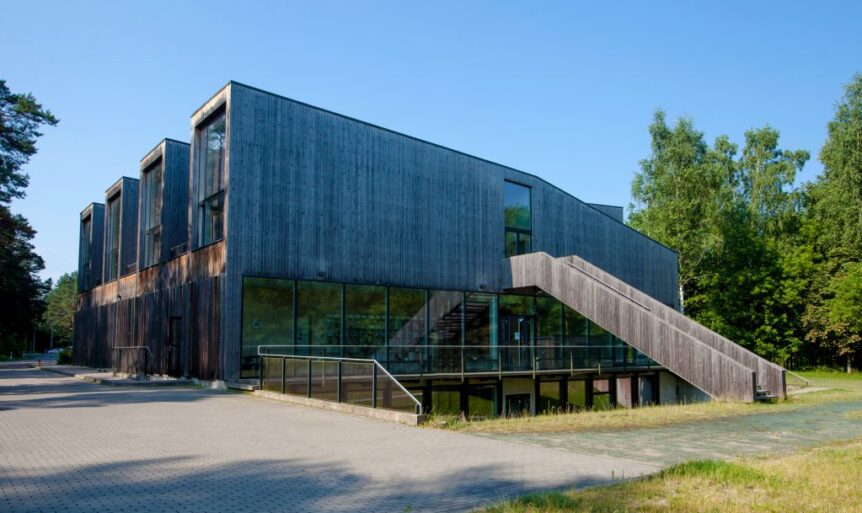 Modern building made primarily of wood and glass is in a green area, perhaps a forest. The weather is nice and sunny.